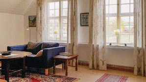 A cozy living room with a blue sofa, a small table, and a lamp. There are large windows with curtains and a framed picture on the wall.