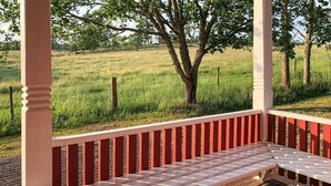 A covered outdoor seating area with a bench and a table, surrounded by trees and a grassy field.