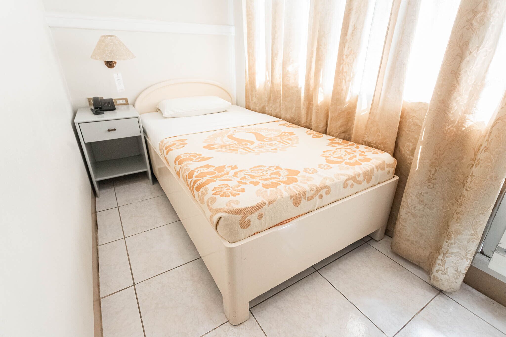 A neatly made bed with a patterned bedspread, a bedside table with a lamp, and a window with curtains.