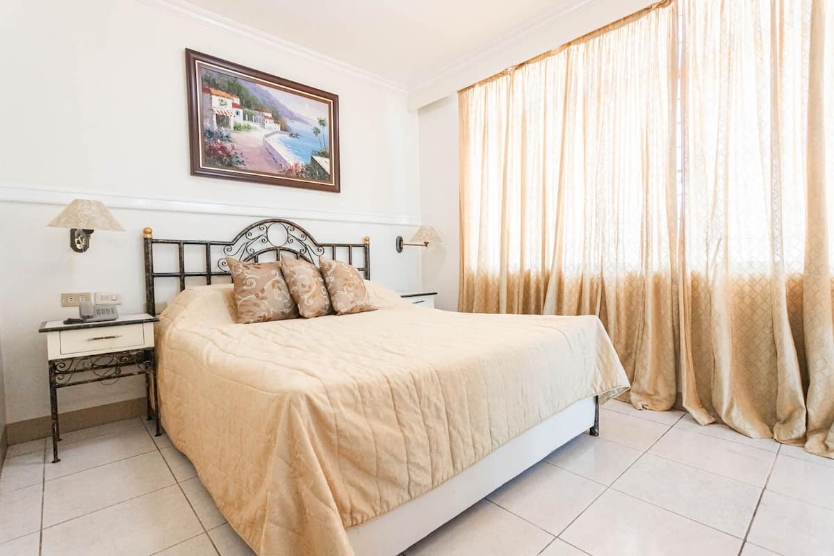 Un dormitorio con una cama, dos mesitas de noche, una lámpara de noche, un cuadro en la pared y una ventana con cortinas.