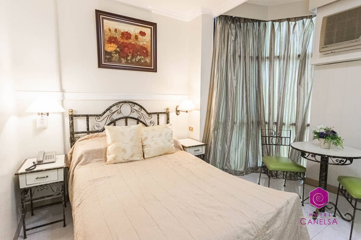 Una habitación de hotel con cama, lámparas de noche, una mesa con sillas, un cuadro en la pared y una cortina.