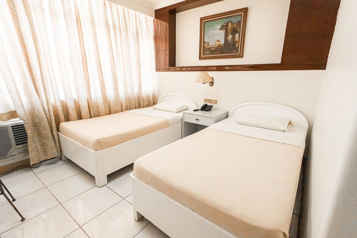 Habitación de hotel con dos camas individuales, una mesita de noche con lámpara, un cuadro en la pared y una ventana con cortinas.