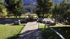 A pathway leading to a seating area with planters and a railing, surrounded by trees and a view of a mountainous landscape. - Hotel Panorama (Cavalese)