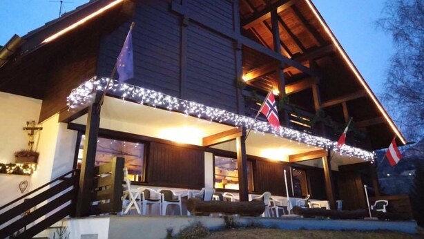 A wooden building with a covered patio area, outdoor seating, and flags. - Hotel Panorama (Cavalese)