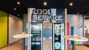 Un quiosco de autoservicio con un rótulo que dice 'Cool Service', máquinas expendedoras y dos mesas con vasos y periódicos.