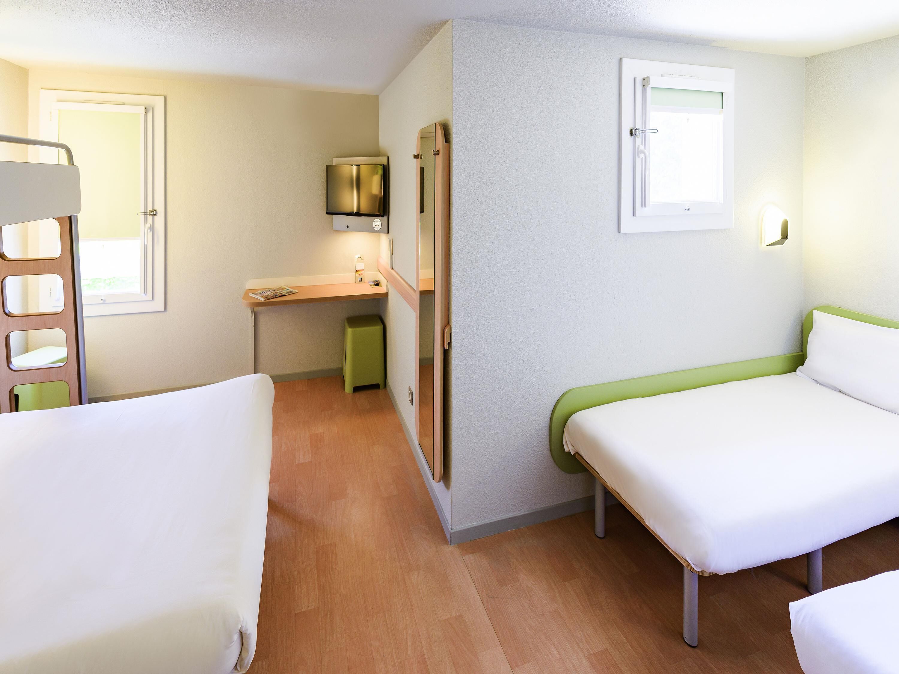A compact hotel room with two single beds, a small desk, a television, and a window.