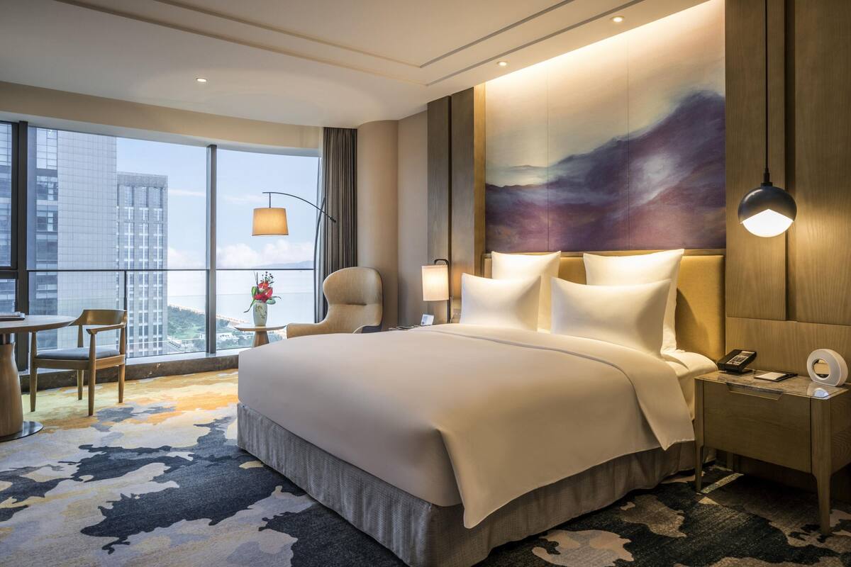 A hotel room with a large bed, a desk with a chair, a nightstand, a lamp, and a painting on the wall.