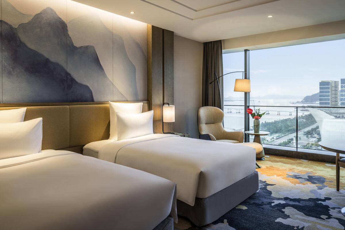 A hotel room with two beds, a large painting, a view of the city, and a balcony with a railing.
