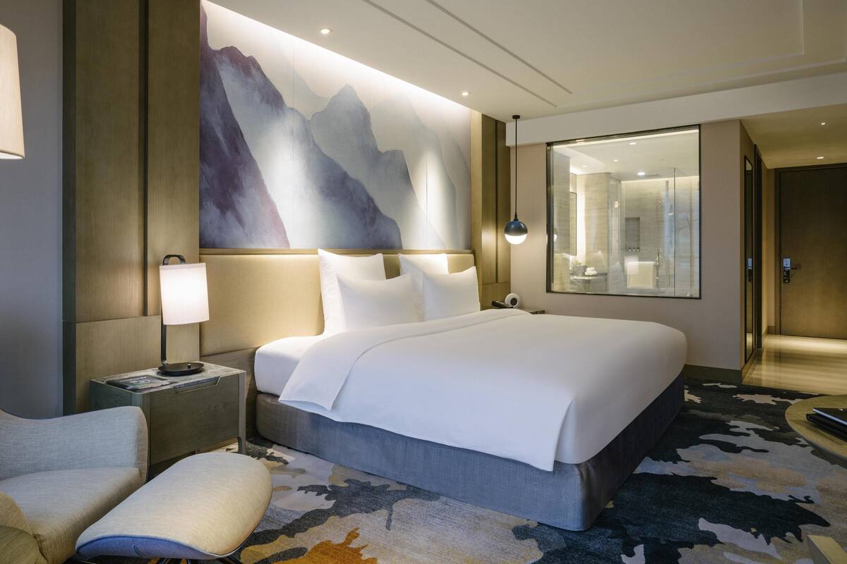 A modern hotel room with a large bed, a nightstand with a lamp, a seating area with a chair and ottoman, and a large mirror on the wall.