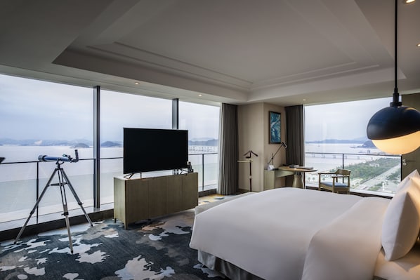 A hotel room with a large bed, a desk with a flat-screen TV, a telescope, and a view of a body of water and cityscape. - Pullman Zhoushan Seaside (Zhoushan)