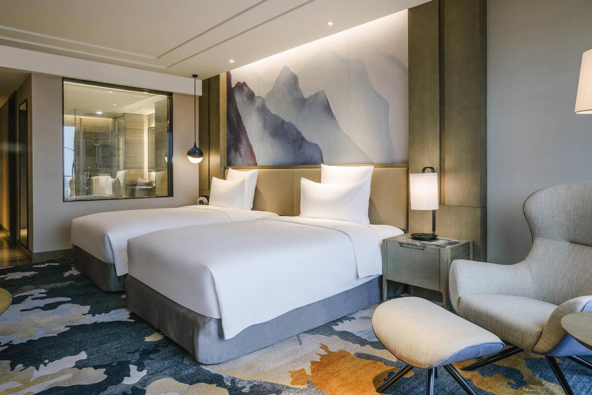 A hotel room with a large bed, a chair, a small table, a mirror, and a wall with a mountain painting.