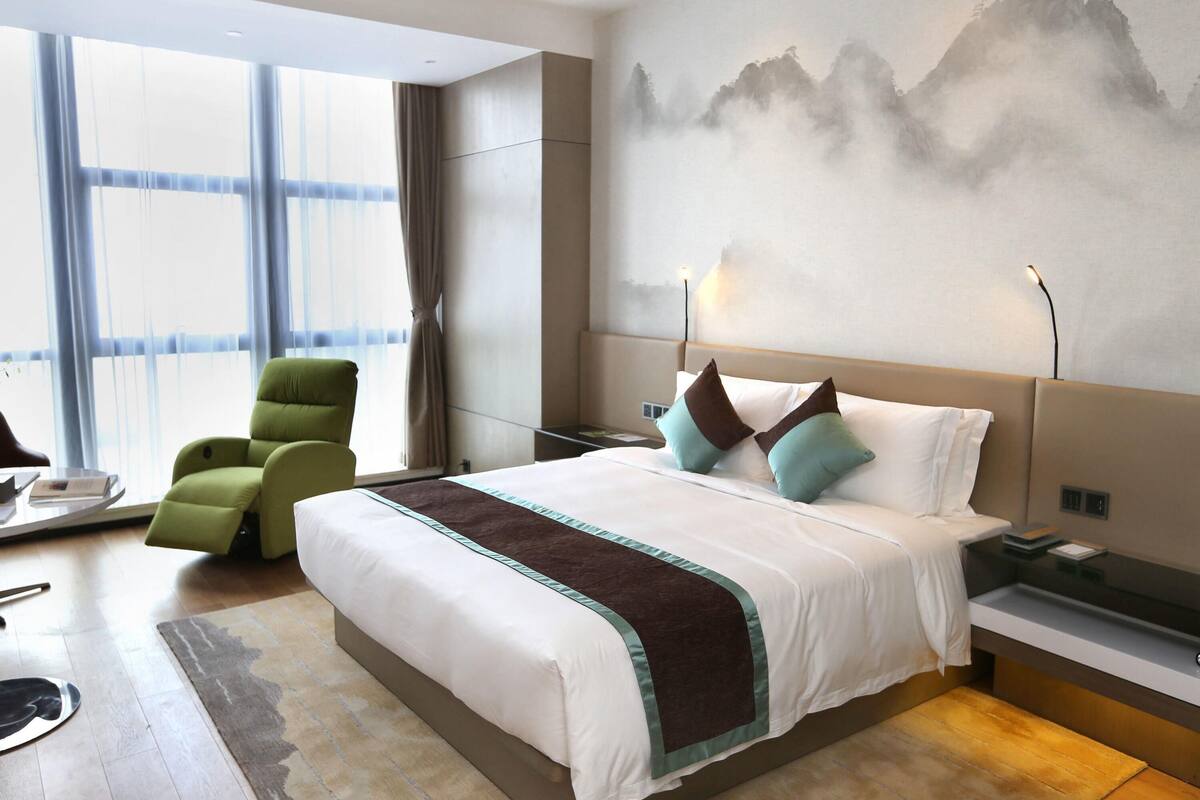 A modern hotel room with a large bed, a green armchair, a view of the city through large windows, and a mountain painting on the wall.