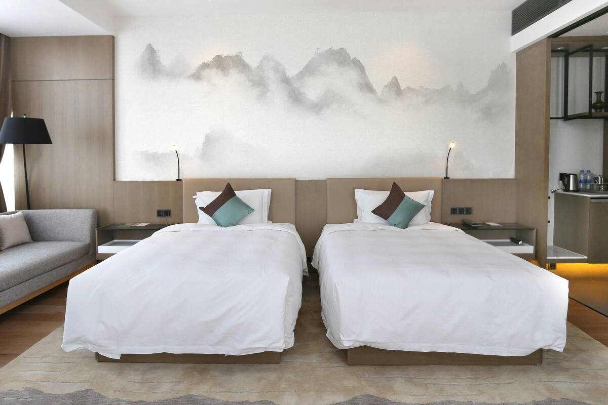A modern hotel room with two beds, a large wall painting, a sofa, and a small kitchenette.