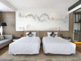 A modern hotel room with two beds, a large wall painting, a sofa, and a small kitchenette.