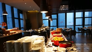 A buffet setup with various food items and utensils, large windows with a city view, and modern lighting fixtures. - Novotel Deqing Moganshan (Huzhou)