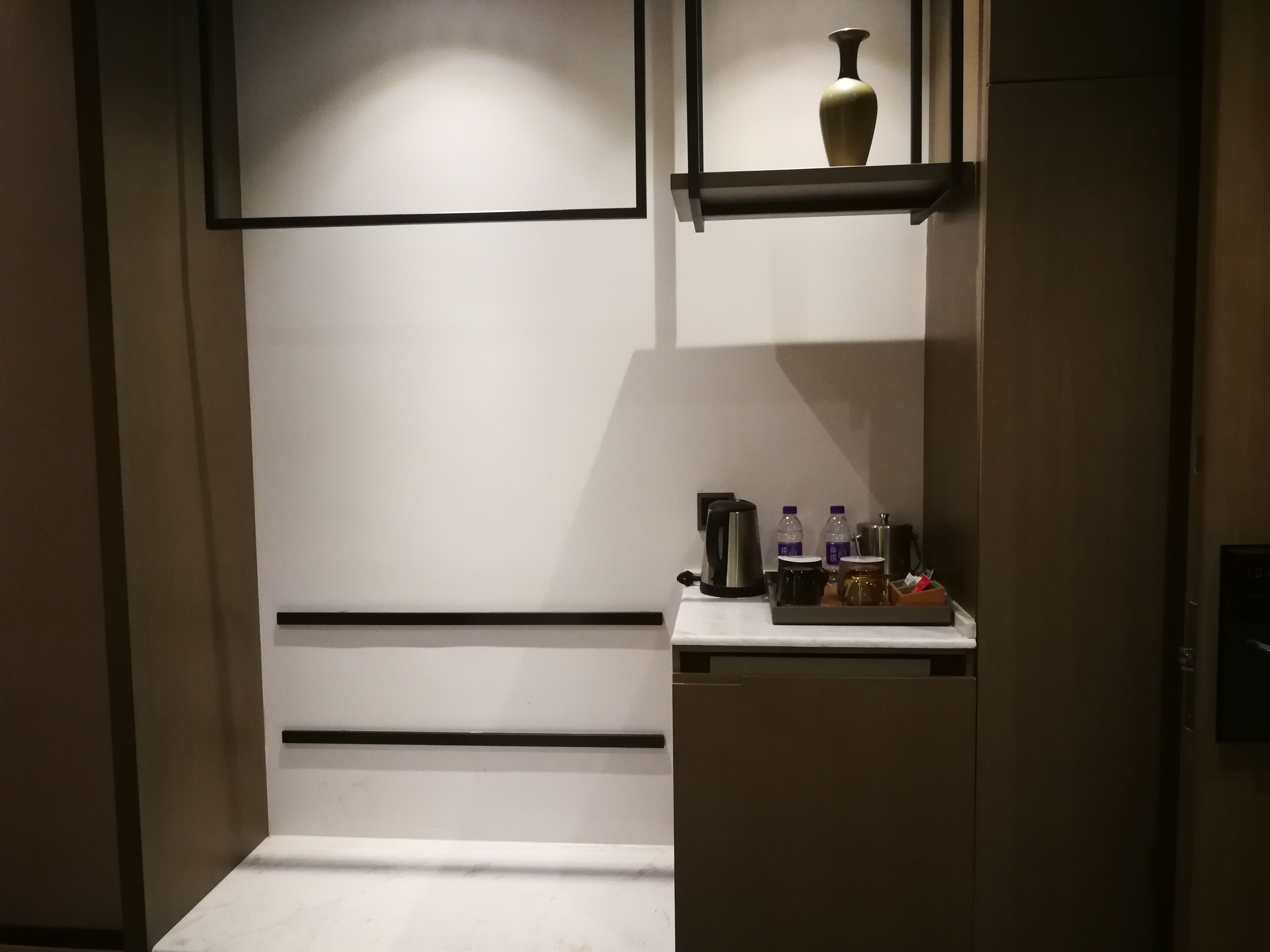 A hotel room with a minimalist design, featuring a white wall, a small shelf with a vase, and a coffee maker.