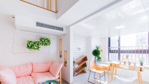 Un salon lumineux avec un canapé rose, une petite table ronde et un bureau avec des chaises. Il y a une fenêtre donnant sur des immeubles et une plante sur le bureau.