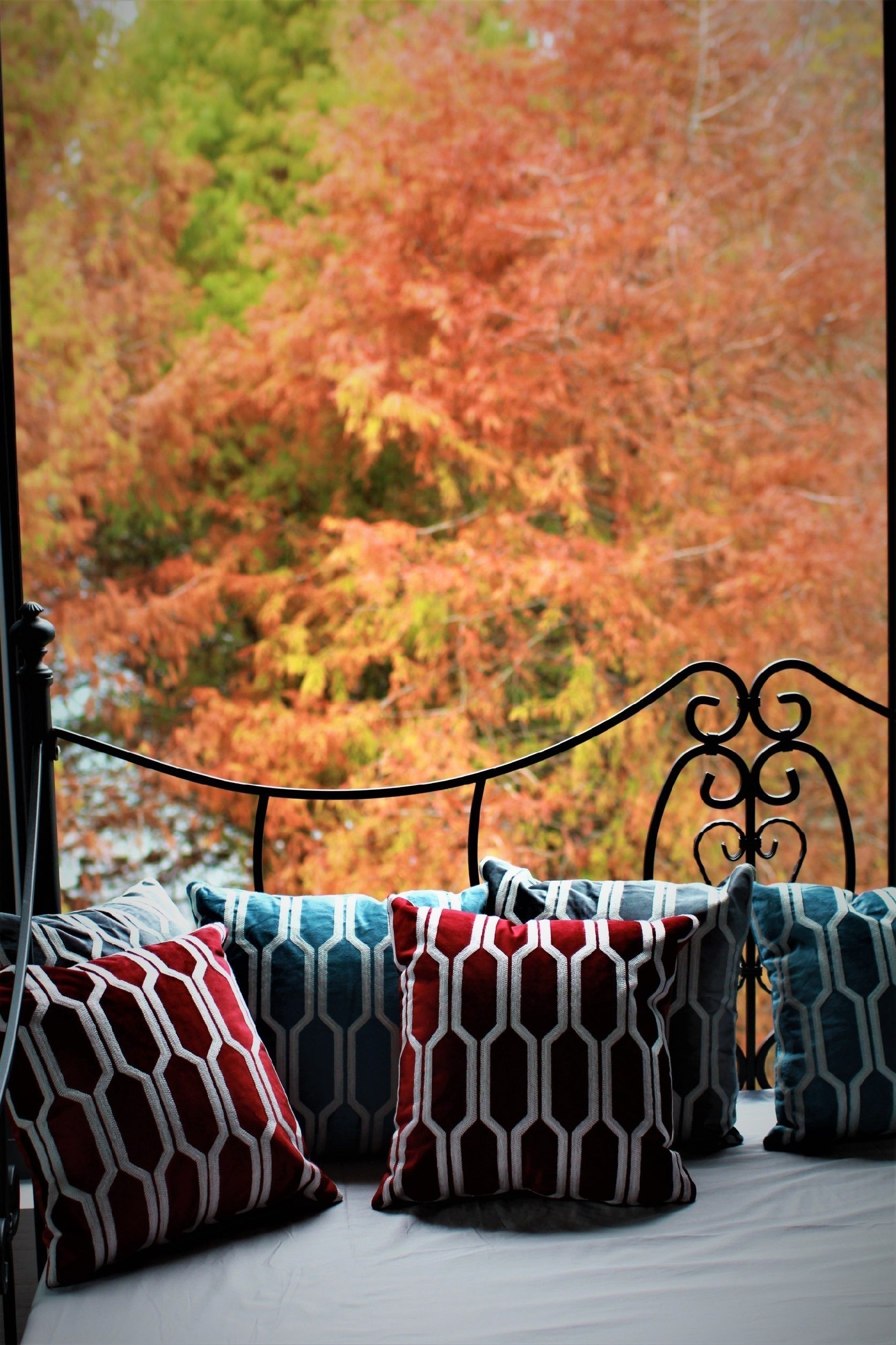A bed with patterned pillows and a metal headboard, set against a backdrop of autumn foliage.