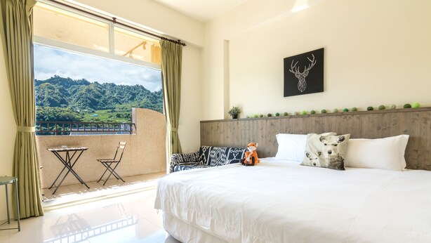 A hotel room with a large bed, a view of a green landscape, a small table and chair, and a decorative deer head on the wall. - Cozy Green Guest House (Taitung)