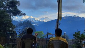 Two people seated facing a mountainous landscape with a wooden fence and greenery in the foreground. - Mochilero Ostello (Gyalshing)