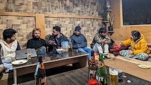 A group of people seated around a wooden table with drinks and plates in a room with a wooden wall pattern. - Mochilero Ostello (Gyalshing)