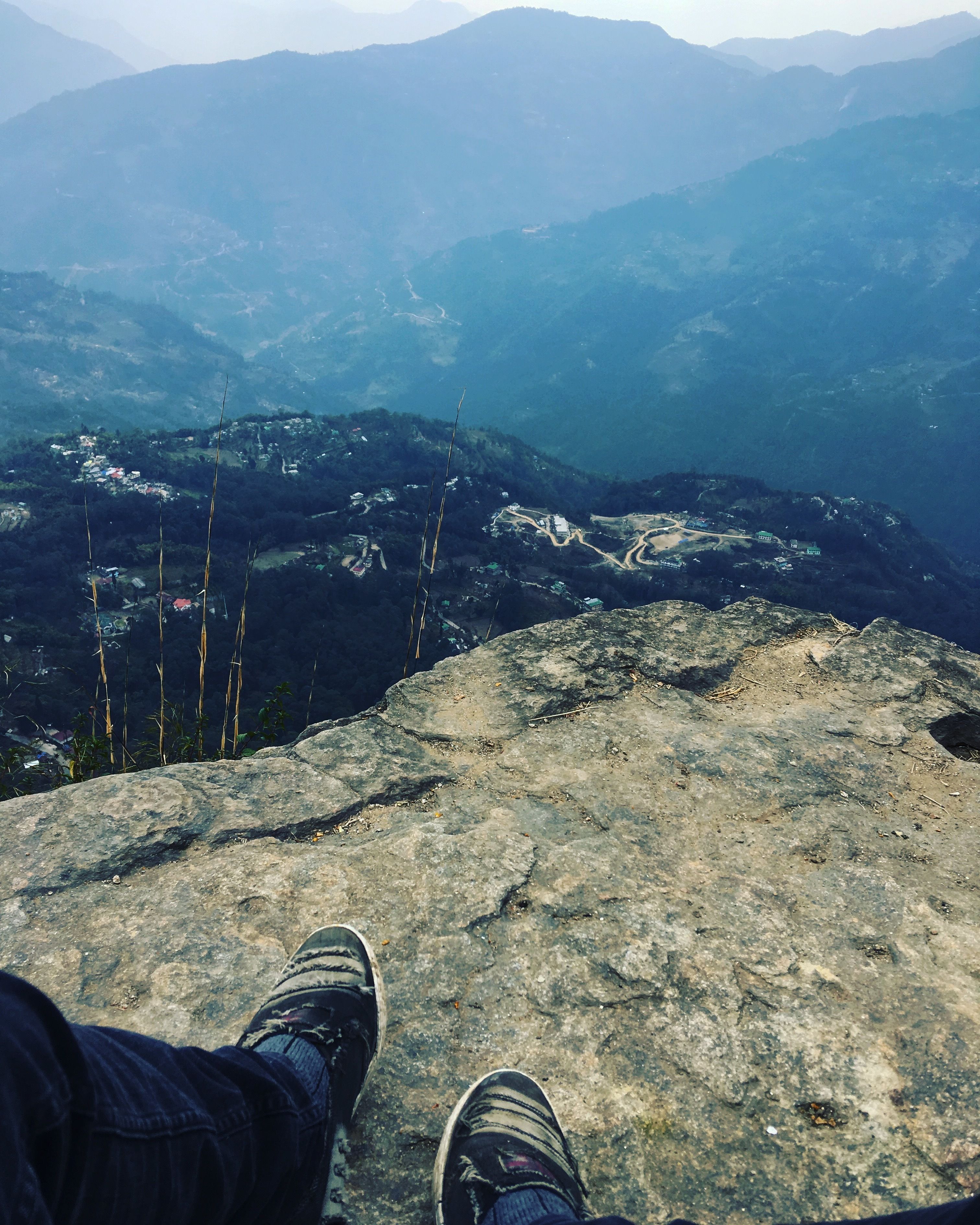 A person's feet on a rocky outcrop overlooking a mountainous landscape with visible roads and buildings.