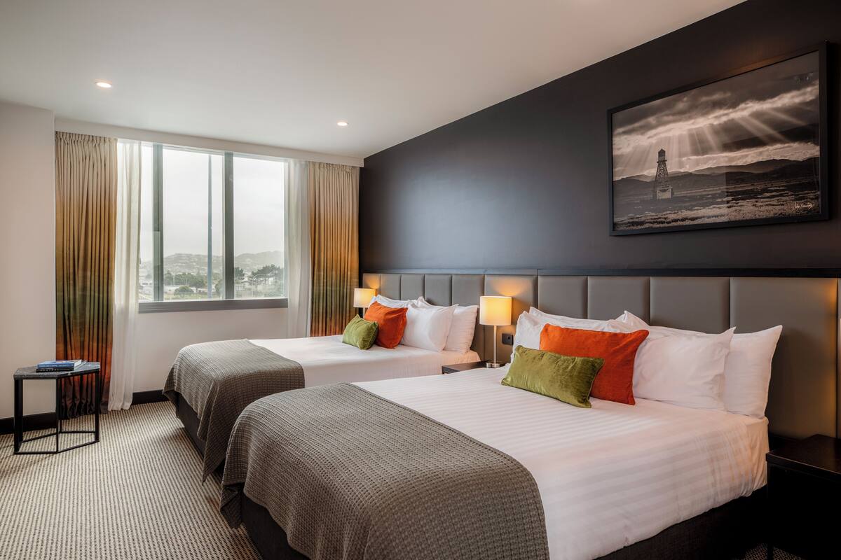A hotel room with two beds, a large window, a painting, and a small table.