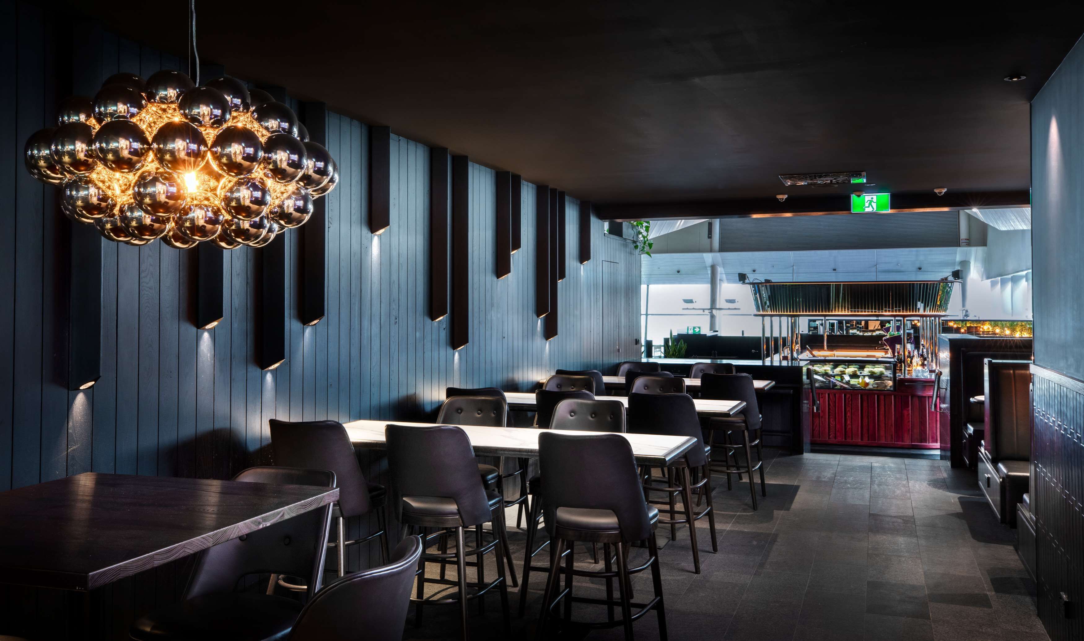 A modern restaurant interior with a unique spherical chandelier, wooden panel walls, and a bar area visible through an open kitchen.