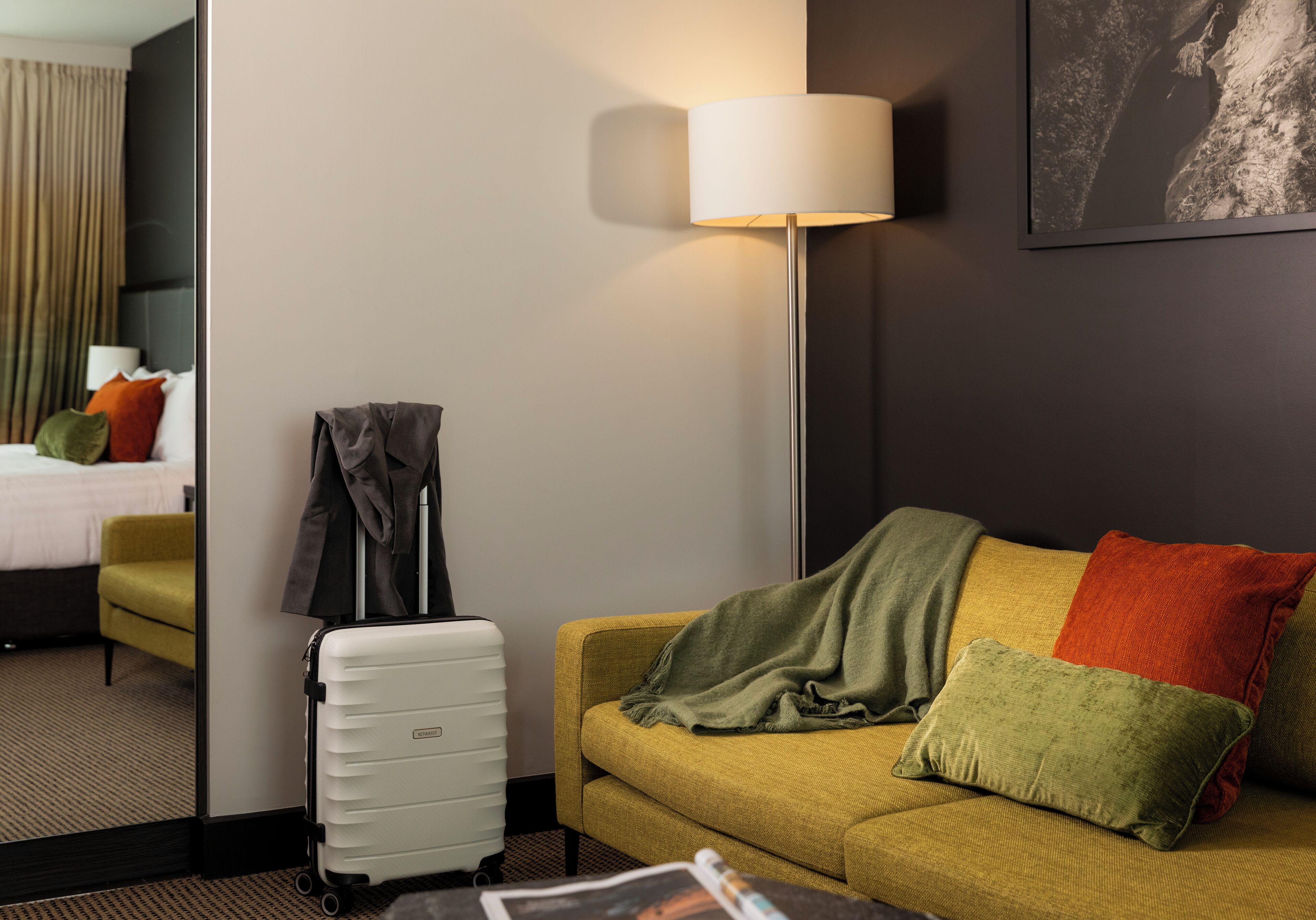 Una habitación de hotel con una cama, un sofá amarillo, un cojín verde y rojo, una maleta blanca y una lámpara.