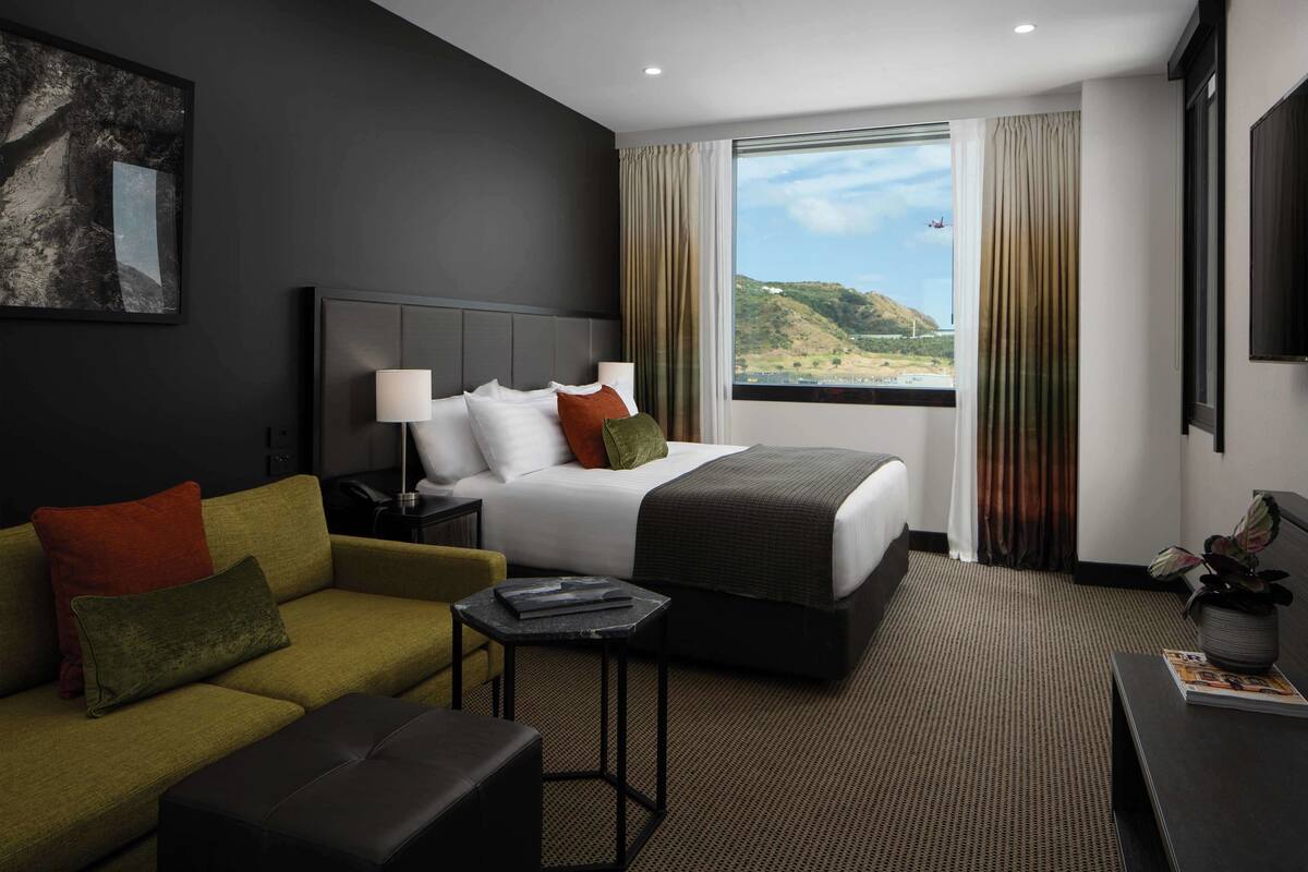 A modern hotel room with a large bed, a sofa, a small table, and a view of the outdoors.