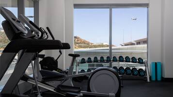 Une salle de sport équipée de divers appareils, un porte-haltères rempli et une vue dégagée sur le ciel et le paysage environnant grâce à de grandes fenêtres.