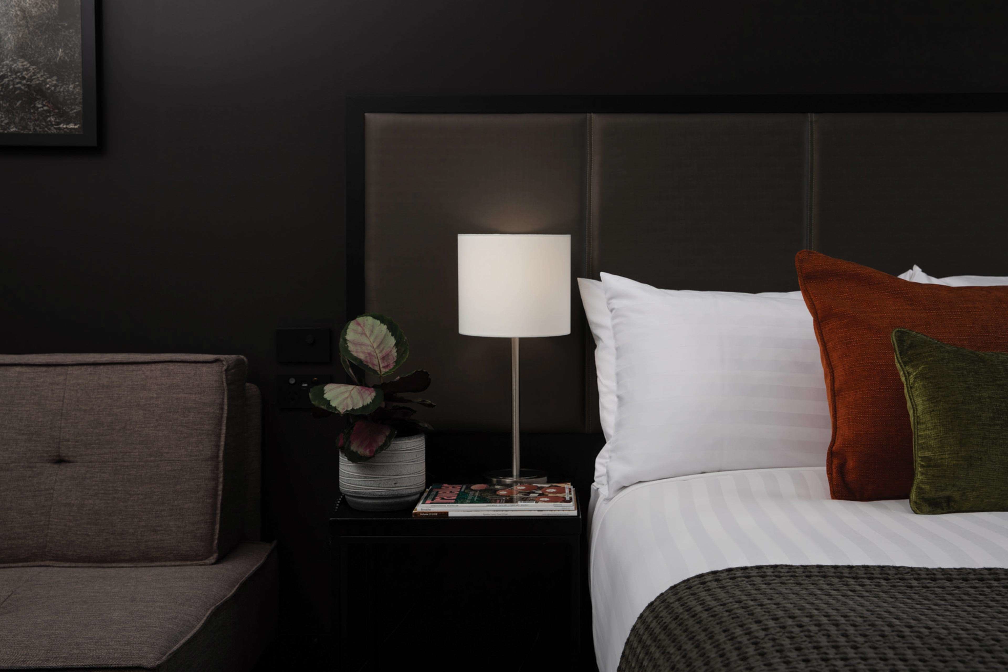 Et pænt indrettet hotelværelse med seng, lille sengebord med lampe, et magasin og en potteplante.