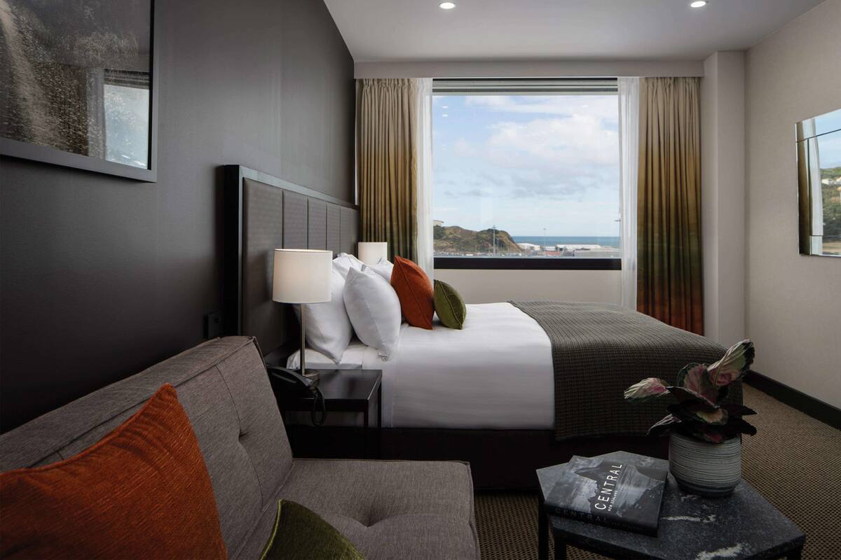 A hotel room with a bed, a sofa, a small table with a book and a plant, and a window with curtains.