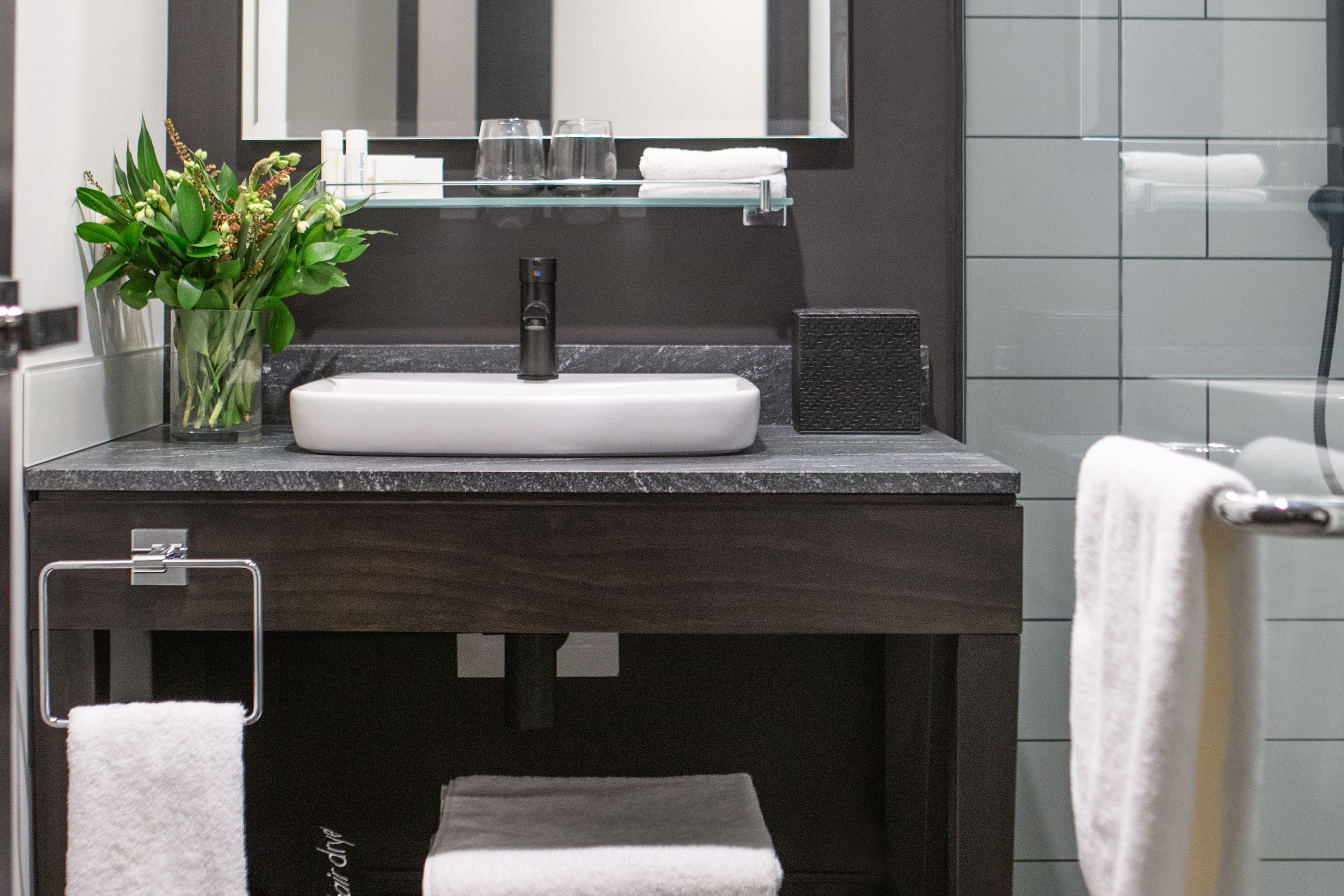 Un baño moderno con un lavabo blanco, encimera oscura y estante de vidrio sobre el lavabo.