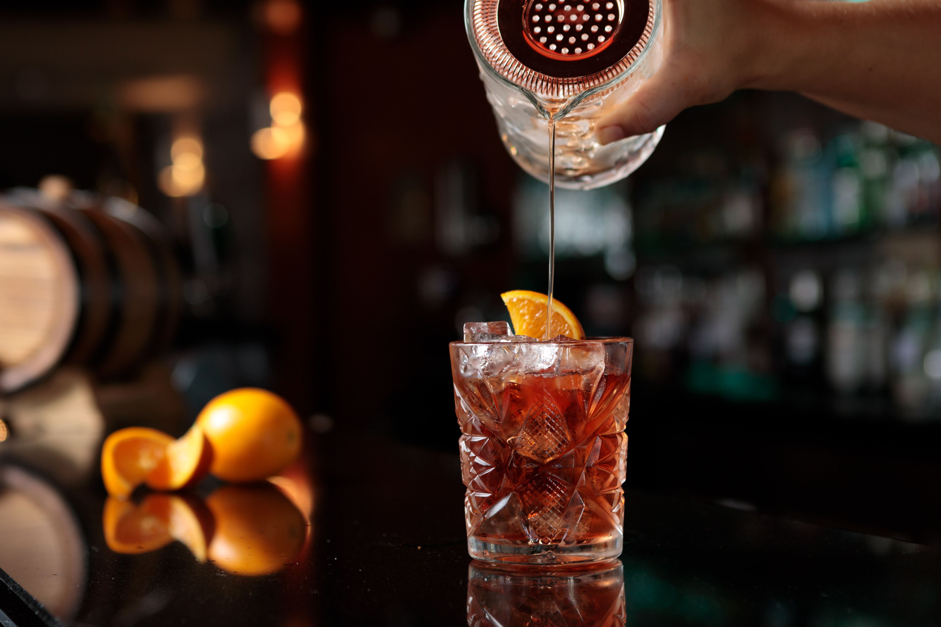 A bar setting with a glass of amber-colored drink garnished with an orange slice, ice cubes, and a bar tool.