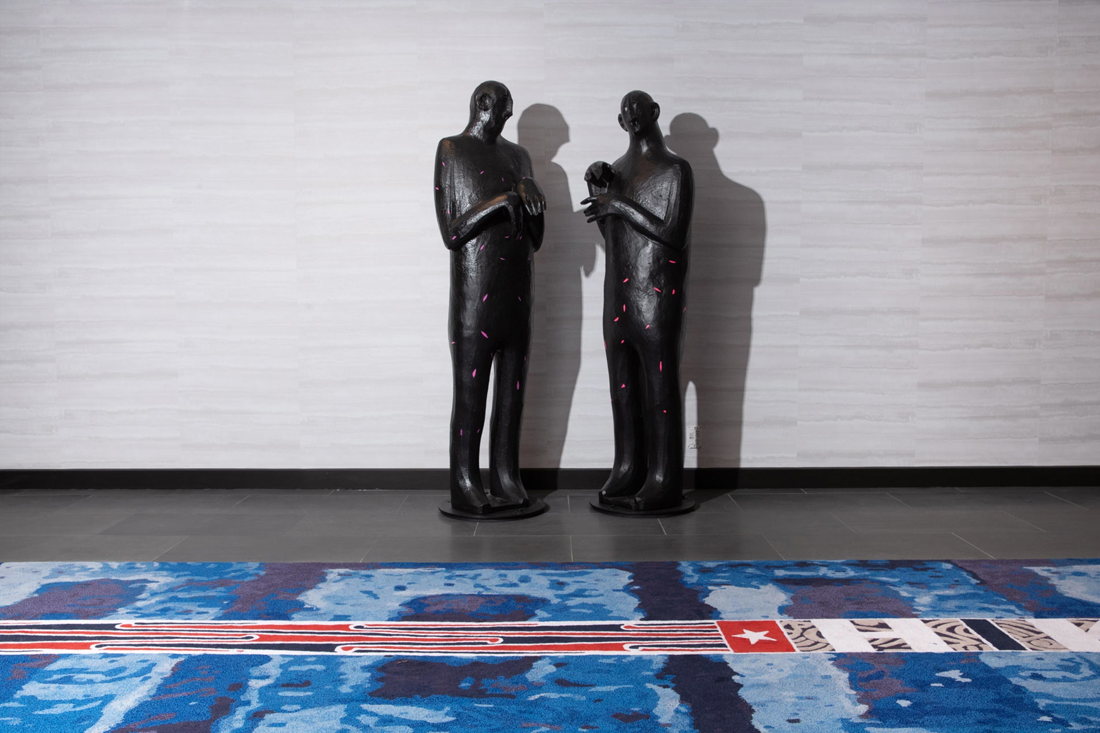 Zwei schwarze Statuen auf einem gemusterten Teppich vor einer schlichten Wand.