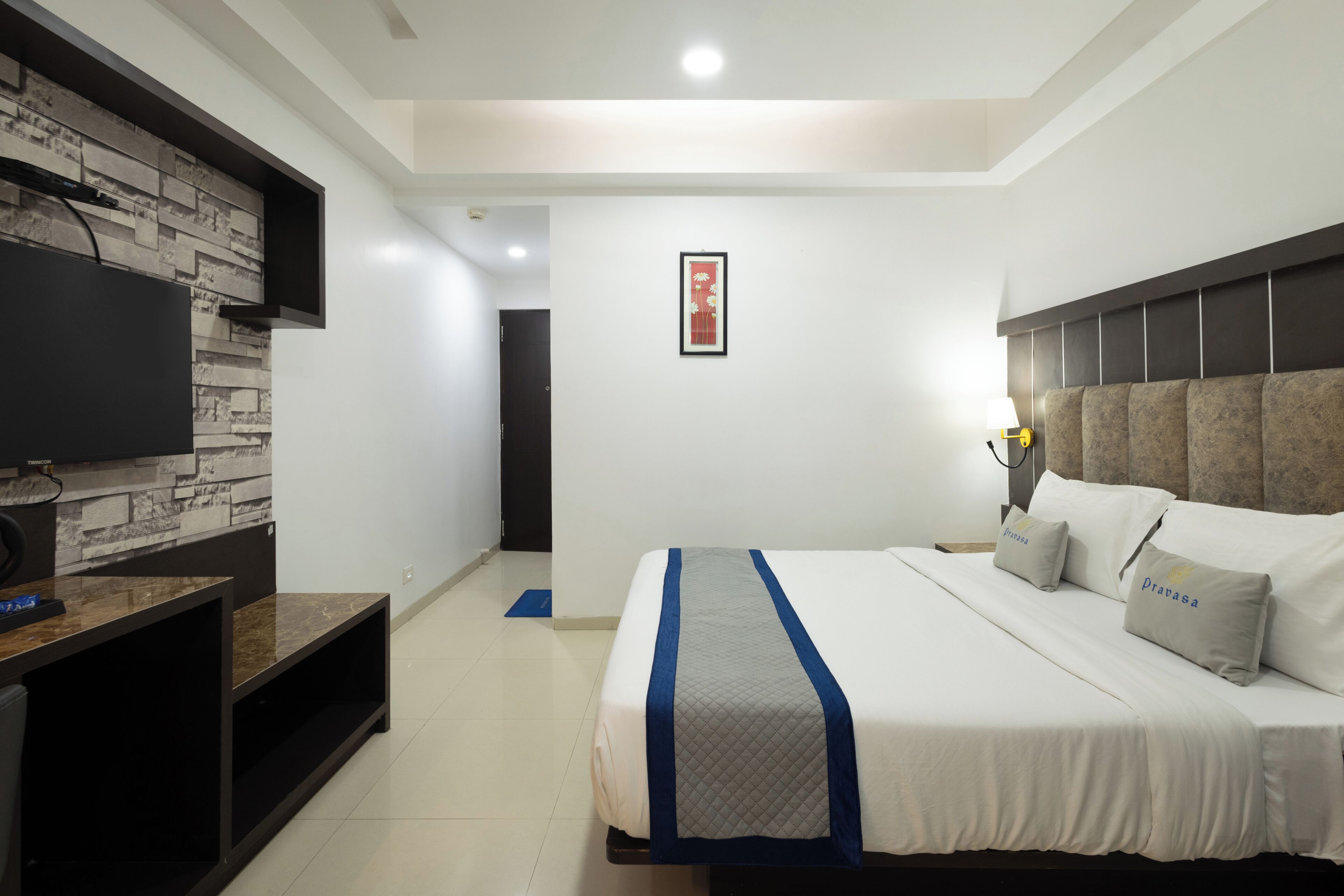 A modern hotel room with a large bed, a flat-screen TV mounted on a stone-textured wall, and a framed artwork above the door.