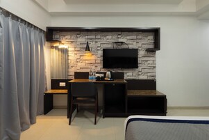 Une chambre d’hôtel compacte comprenant un lit, un bureau, une chaise, un téléviseur fixé à un mur à l’aspect pierre, et une fenêtre avec des rideaux.