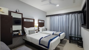 A hotel room with two beds, a desk with a kettle, a mirror, a ceiling fan, and a window with curtains. - Pravasa Hotel by Sapphire, Vadodara (Vadodara)