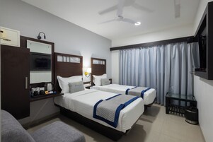 Habitación de hotel con dos camas, un escritorio con hervidor, un espejo, ventilador de techo y ventana con cortinas.