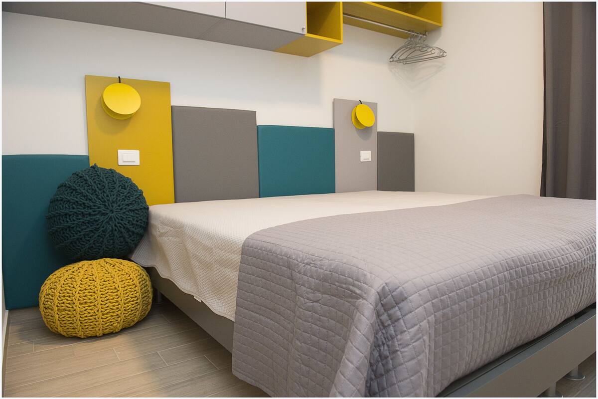 Une chambre à coucher moderne avec un lit, un mur d’accent aux couleurs jaune et turquoise, et un grand pouf tricoté.