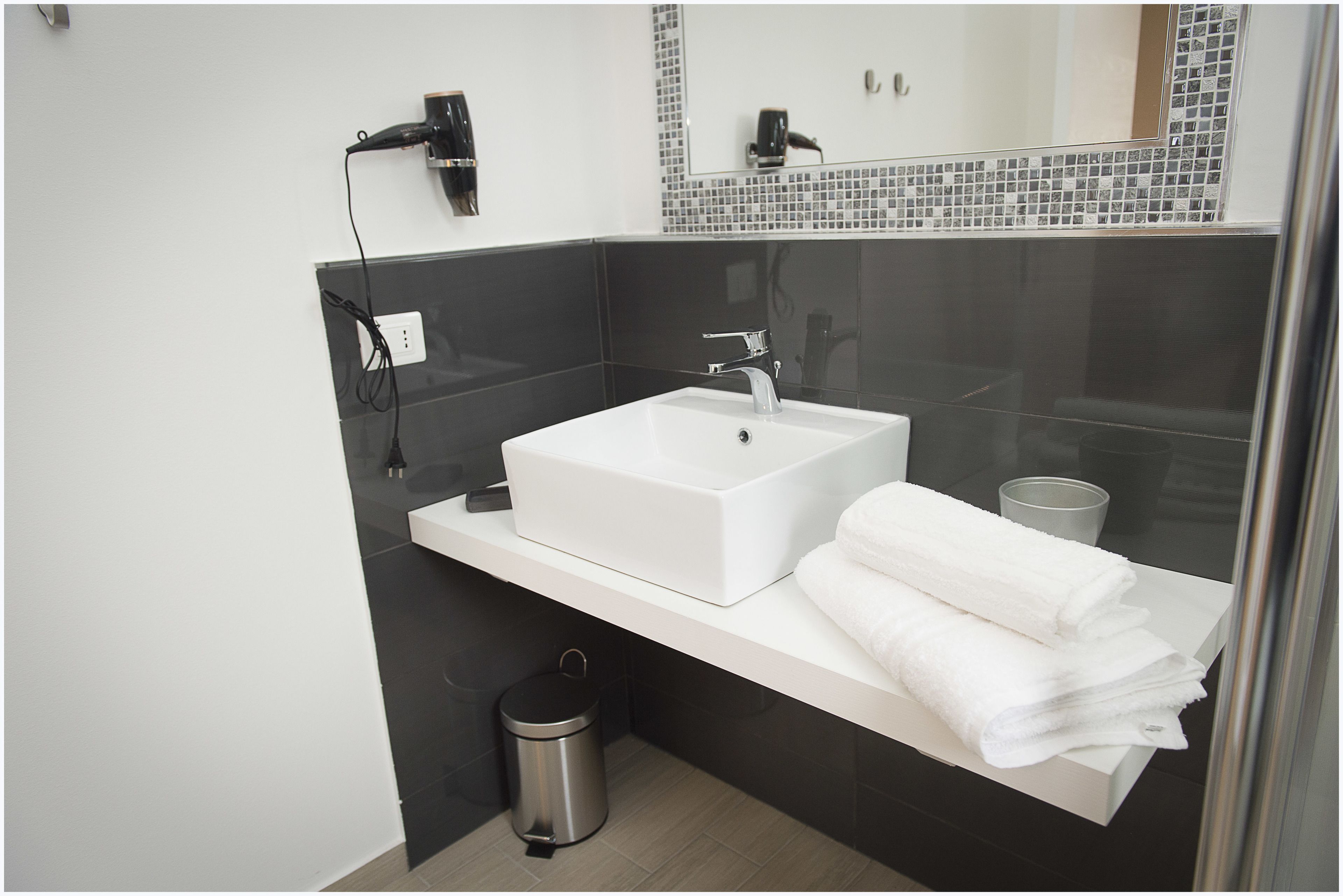 Une salle de bain moderne avec un lavabo blanc, des carreaux noirs, un miroir avec un pare-feu en mosaïque, un porte-serviettes et une poubelle.
