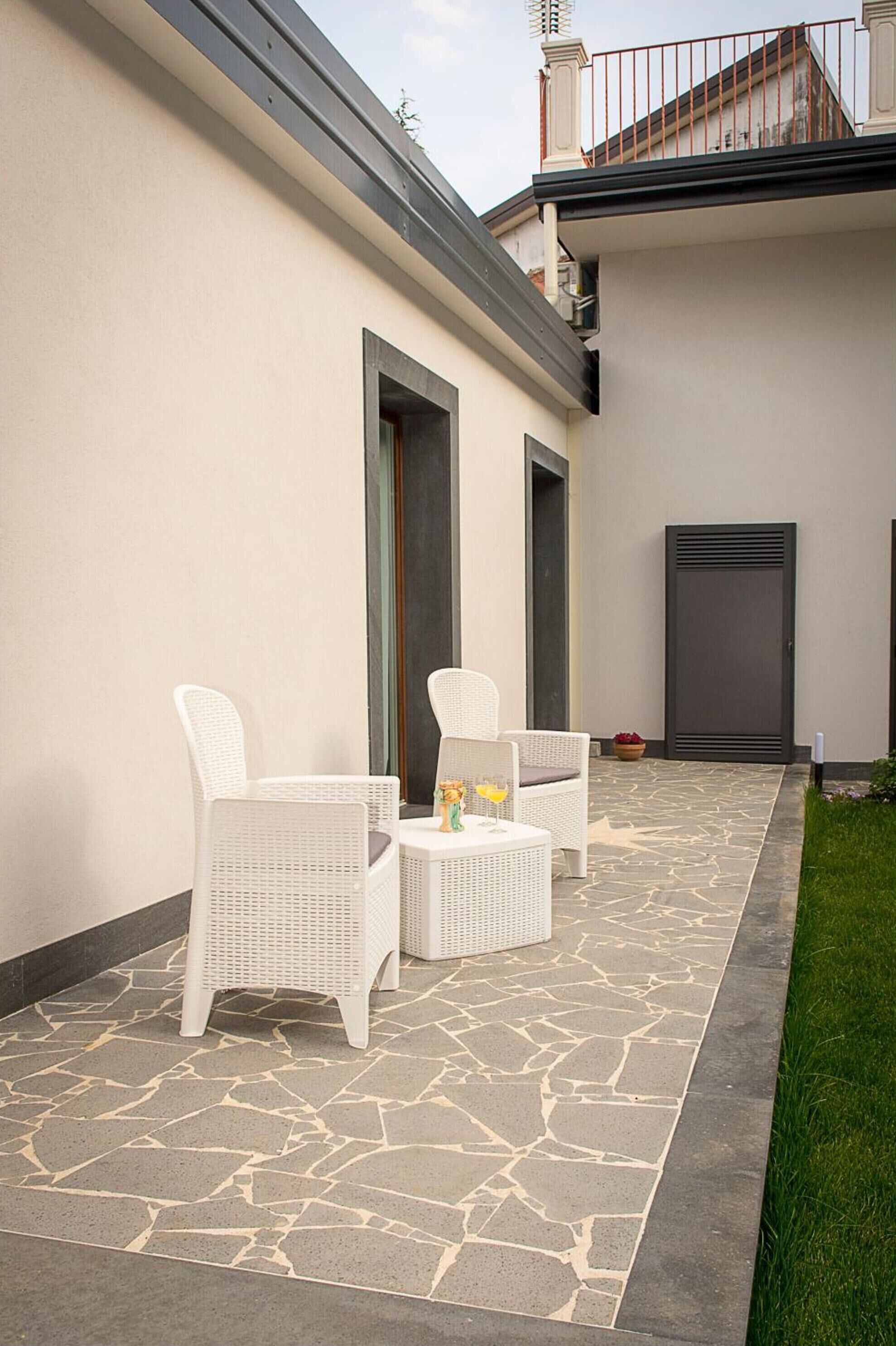 A patio with two white wicker chairs and a small table, a stone-tiled floor, and a white wall with a door and a window.