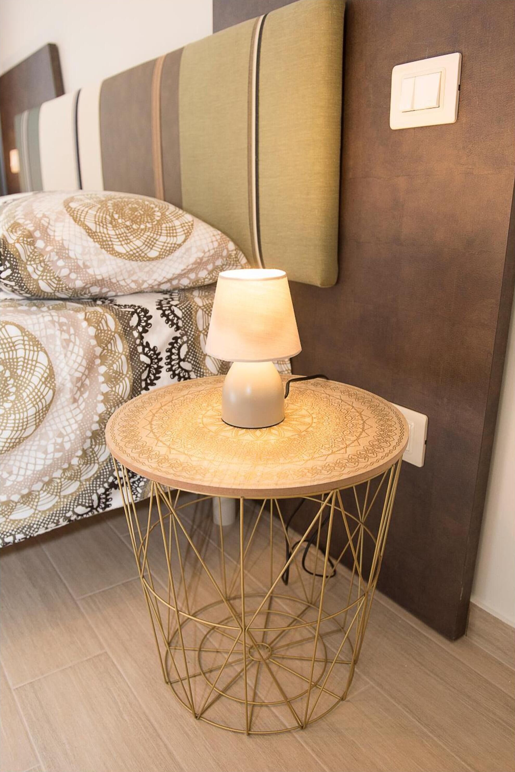 A bed with patterned bedding, a bedside lamp on a round table, and a wall switch.