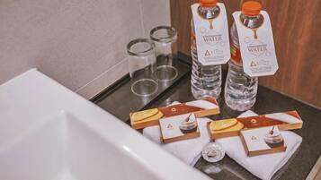 Un meuble-lavabo de salle de bain avec deux bouteilles d'« eau offerte » de la marque Azana, deux récipients en verre vides et deux boîtes de « gel douche Azana ».