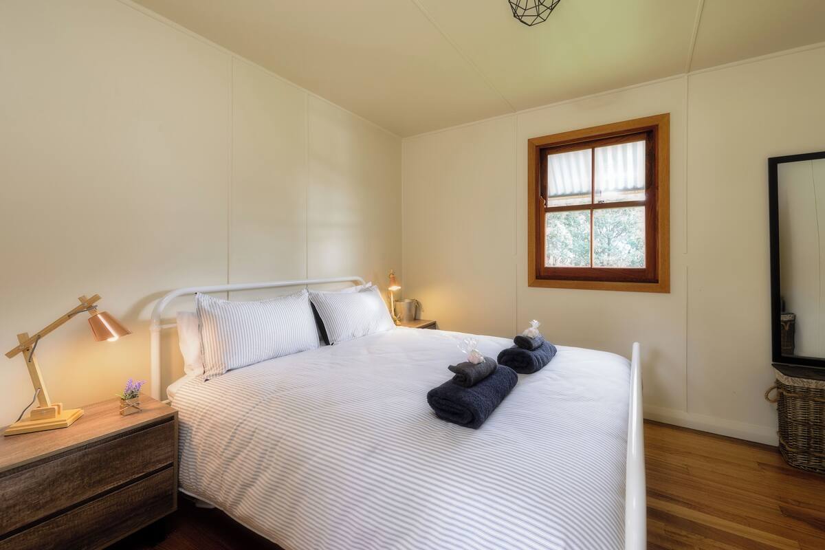 Una cama bien hecha con ropa de cama a rayas, una lámpara de noche y una ventana con vistas a árboles.
