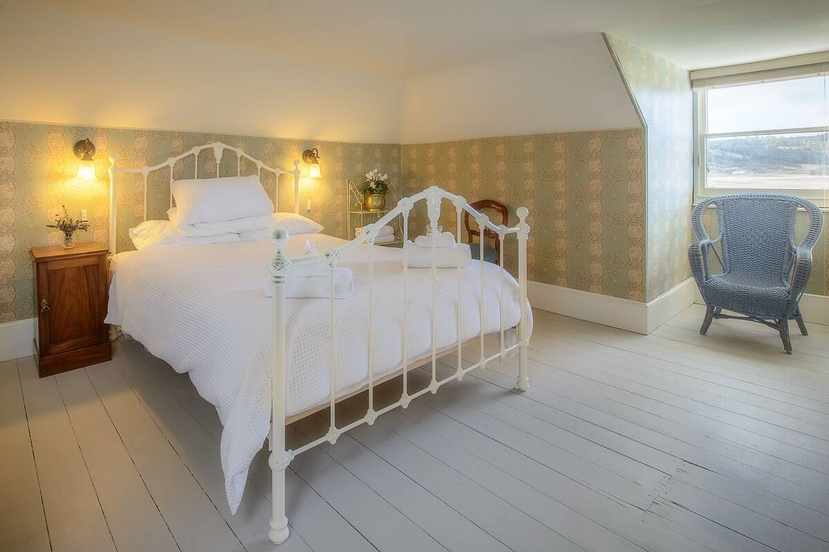 Un dormitorio con una cama de metal blanco, dos lámparas de noche, una mesita de noche de madera y una silla de mimbre.