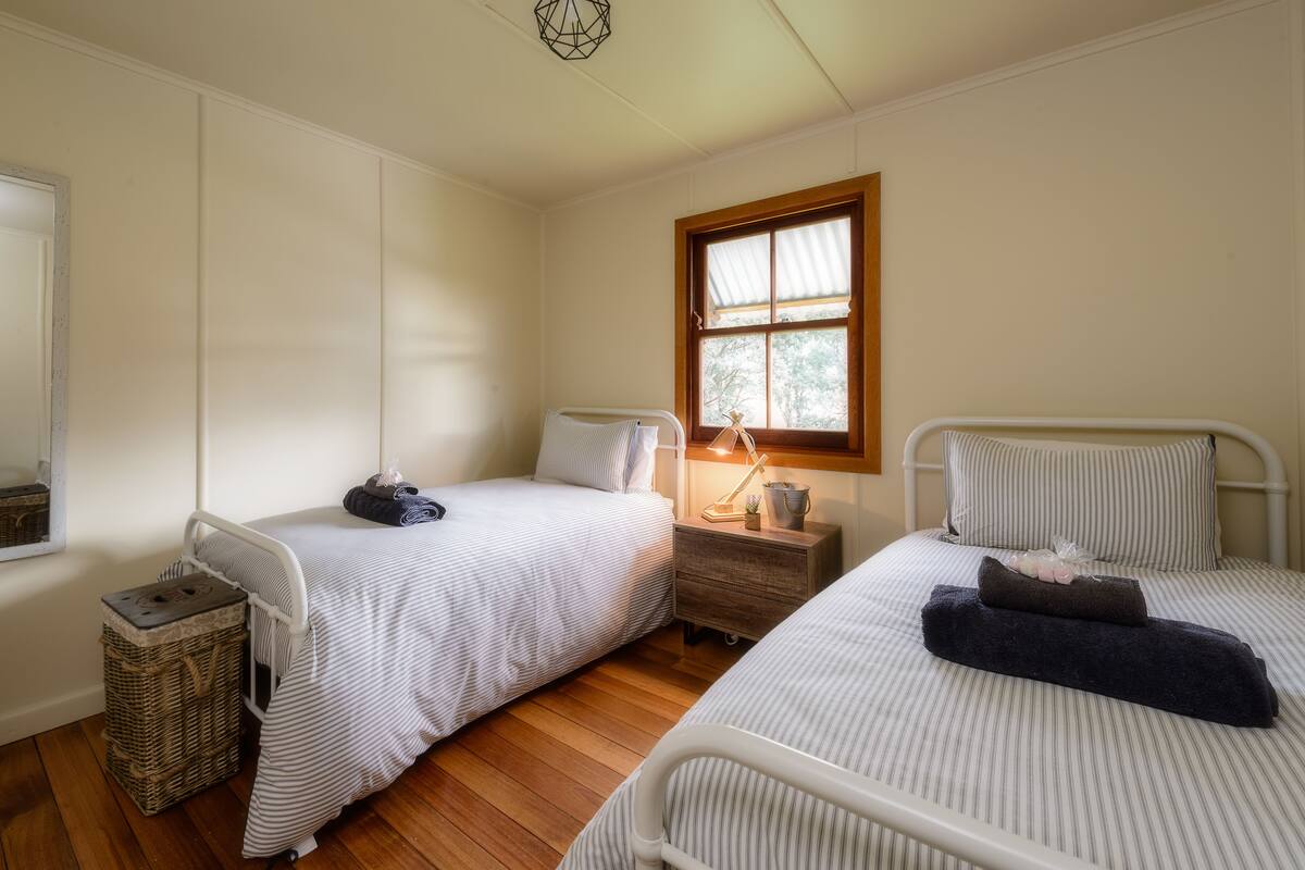 Un dormitorio con dos camas individuales, piso de madera, ventana con marco de madera, mesita de noche con lámpara y una pequeña cesta de mimbre.