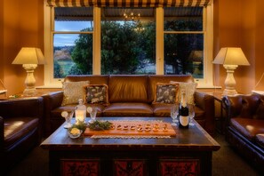 A cozy living room with a brown leather sofa, two side tables with lamps, a wooden coffee table with a decorative tray, and a large window with a view of greenery.