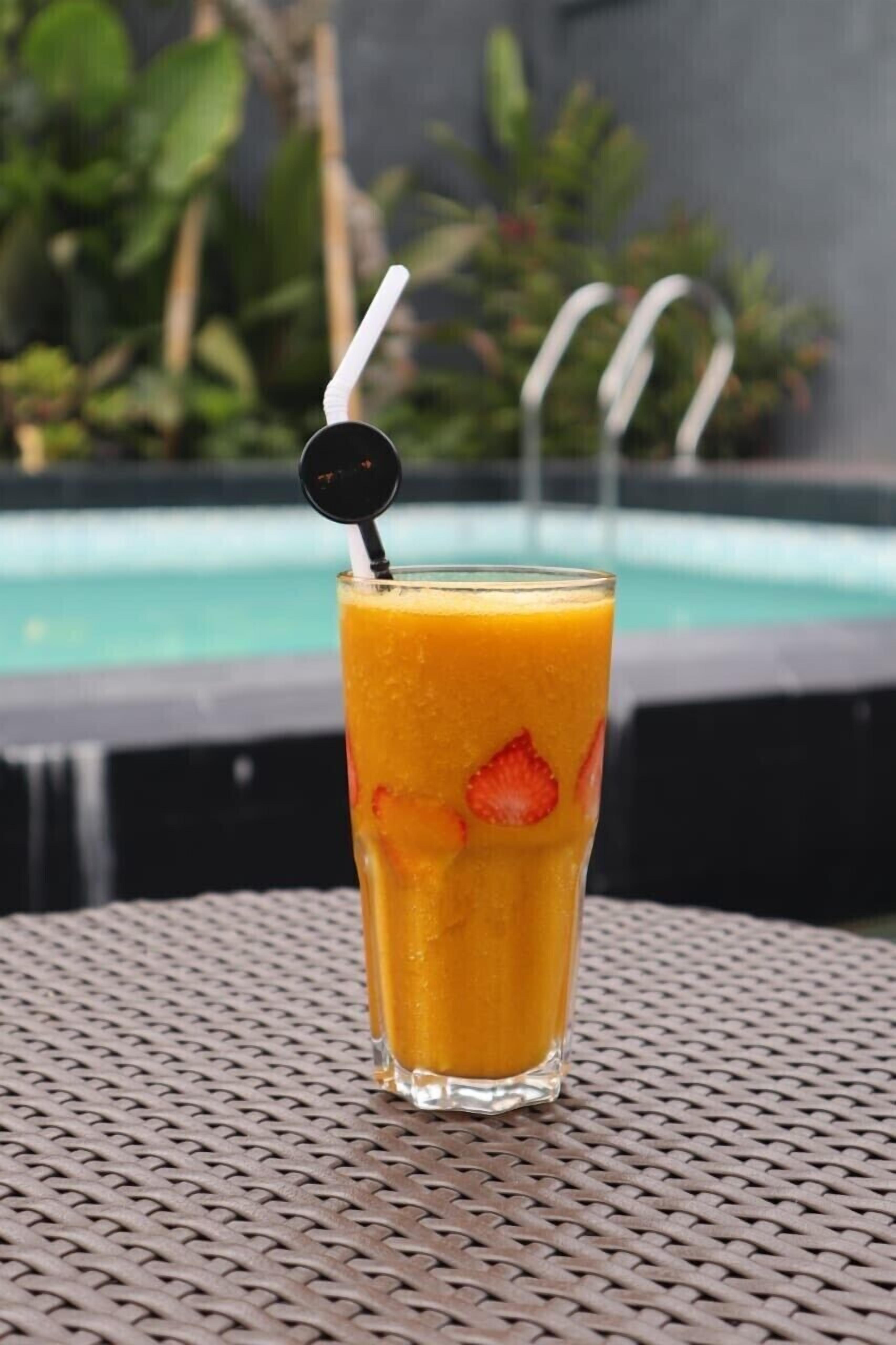 A tall glass of orange juice with a straw and a black cap, placed on a woven table with a pool and greenery in the background.
